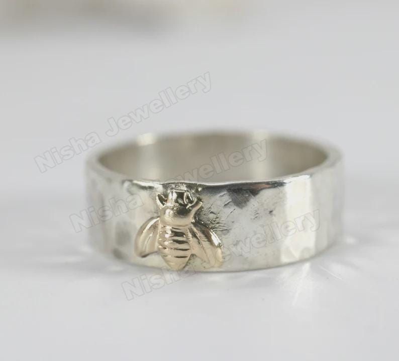 May include: A silver ring with a hammered texture and a gold-colored bee charm. The bee is centered on the ring and has detailed wings and body. The ring is a wide band style.