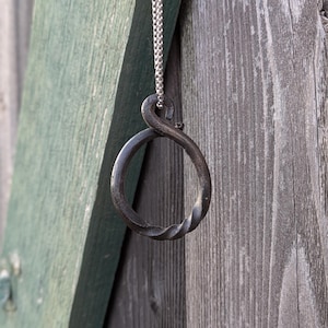 May include: A handcrafted, dark metal pendant necklace. The pendant features a twisted, circular design, suspended from a delicate silver chain. The necklace hangs against a weathered wooden background, with a green painted wooden plank visible.