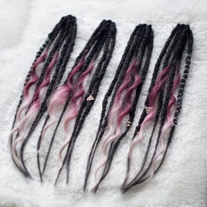 May include: Four black dreadlocks with pink and white ombre accents. Each dreadlock features a unique design, with some incorporating small gold-colored charms and beads. The dreadlocks are arranged on a white, textured surface.