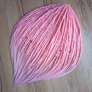 May include: A bundle of pink synthetic dreadlocks with a light pink ombre effect. The dreads are adorned with small silver and red beads. The dreadlocks are arranged on a wooden surface.