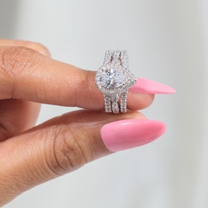 以下が含まれることがあります： ハローセッティングと小さなダイヤモンドのバンドが付いた、洋梨型のダイヤモンドの婚約指輪。指輪はシルバー製で、ピンクのネイルポリッシュを塗った指につけられています。