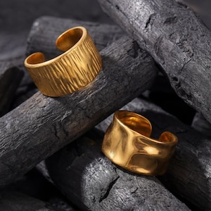 May include: Two gold-colored adjustable rings. One ring has a textured surface, while the other has a smooth, hammered appearance. The rings are displayed on a bed of dark charcoal, creating a contrast in textures and colors. Jewelry.