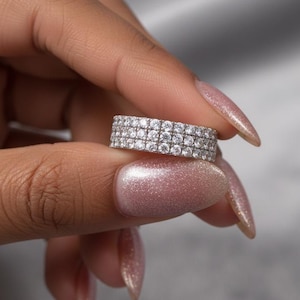 Könnte beinhalten: Ein silberner Ring mit drei Reihen kleiner, klarer Edelsteine. Der Ring wird zwischen Fingern vor einem weichen, verschwommenen Hintergrund gehalten. Der Ring ist im Bandstil gehalten, mit eng aneinander liegenden Edelsteinen.