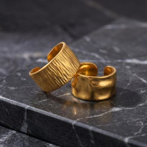 May include: Two gold-colored adjustable rings. One ring has a textured, ridged surface, while the other has a smooth, hammered appearance. The rings are displayed on a dark gray marble surface, showcasing their metallic shine.