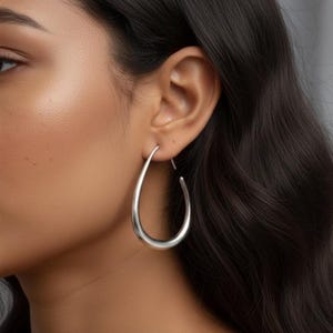 Puede incluir: Pendientes de aro plateados con forma de lágrima. Estos pendientes tienen un acabado liso y pulido y un diseño sencillo y elegante. Los aros son de tamaño mediano, adecuados para el uso diario o para ocasiones especiales. Los pendientes se muestran en la oreja de una persona.