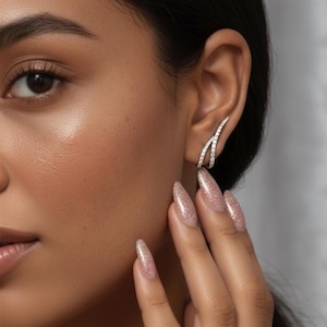 May include: A pair of silver-toned ear cuffs with a curved design, encrusted with small, sparkling stones. The earrings are worn on the ear, and the image shows a close-up of the ear and part of the face.