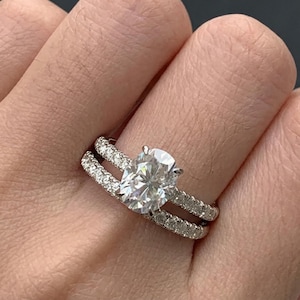 May include: A diamond engagement ring with a large oval-cut center stone and a double band. The bands are encrusted with smaller diamonds, creating a sparkling effect. The ring is set in a silver or white gold setting.