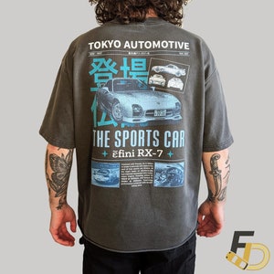 Può includere: Una t-shirt color antracite con una stampa grafica in stile vintage che presenta un'illustrazione blu e bianca di un'auto sportiva. Il testo sulla t-shirt recita "TOKYO AUTOMOTIVE", "登場", "THE SPORTS CAR" e "ɛfini RX-7".