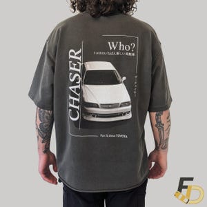 May include: Dark grey t-shirt with a white car graphic. The shirt displays the word "CHASER" vertically on the left and "Who?" with Japanese text above the car. The text "Fun To Drive TOYOTA" is at the bottom.