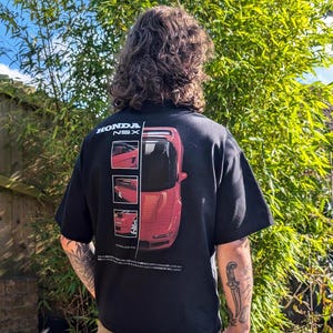 Op de afbeelding: Zwart T-shirt met een rode Honda NSX auto-afbeelding op de rug. De afbeelding toont meerdere afbeeldingen van de auto vanuit verschillende hoeken. De tekst "HONDA NSX" is in wit boven de afbeelding gedrukt.