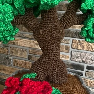 May include: A handmade crochet tree with a brown trunk and green foliage. Red crochet flowers and green leaves are at the base of the tree. The tree is set in a brown pot.
