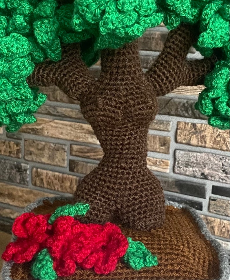 May include: A handmade crochet tree with a brown trunk and green foliage. Red crochet flowers and green leaves are at the base of the tree. The tree is set in a brown pot.