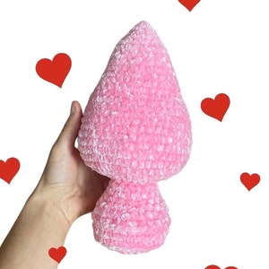 Puede incluir: Un juguete de peluche de hongo de crochet rosa. El hongo es suave y peludo y tiene una parte superior redondeada y un tallo.