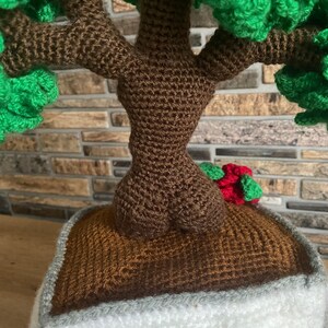 May include: A crocheted bonsai tree with a brown trunk and green foliage. The tree is planted in a brown pot with a white and grey border. A small red and green accent is visible.