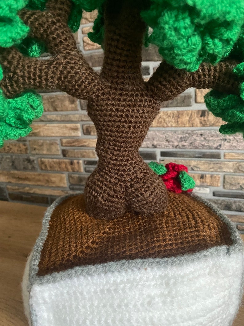 May include: A crocheted bonsai tree with a brown trunk and green foliage. The tree is planted in a brown pot with a white and grey border. A small red and green accent is visible.