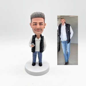 May include: A custom figurine of a person with gray hair wearing a white shirt, black vest, and blue jeans. The figurine is holding a small gray object in their right hand.