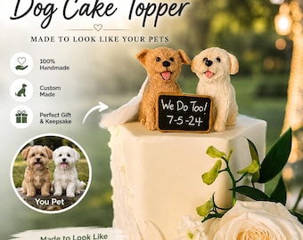 May include: A handcrafted dog cake topper featuring two small dog figurines, one light brown and one cream, sitting on a white cake. The dogs hold a small black sign with the white text We Do Too! 7-5-24. A white veil is attached to the brown dog.