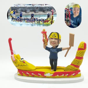 May include: A figurine of a person in a blue shirt and black trousers, holding a wooden paddle and a trophy. The figurine is on a yellow and red dragon boat. The image includes a photo of a dragon boat race and a person holding a trophy.