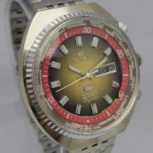 Peut inclure: Une montre Omax vintage avec un bracelet et un boîtier en métal argenté. Le cadran de la montre est un dégradé d'or et de noir, avec une lunette rouge indiquant les noms des villes du monde. La montre affiche le jour et la date.