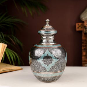 Puede incluir: Una urna decorativa plateada y azul con intrincados patrones florales y una tapa. La urna tiene una banda de color cobre alrededor del medio.