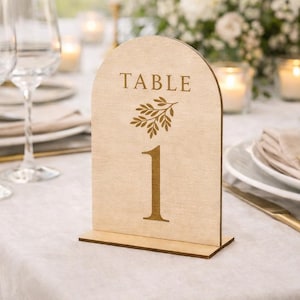 Peut inclure: Un numéro de table en bois avec un sommet en forme d'arche. Le mot "TABLE" est imprimé en haut, avec une branche de feuilles en dessous. Le numéro "1" est imprimé en gros caractères. Le numéro de table est en bois clair.