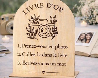 May include: A wooden sign with the words "LIVRE D'OR" at the top, and instructions below in French. The sign features a camera illustration and is designed for a guest book at a wedding or event.