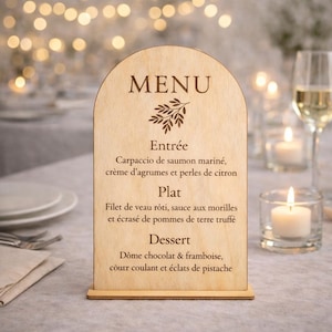 Peut inclure: Un menu en bois avec un dessus en forme d'arche, affichant un menu avec du texte en français. Le menu comprend les plats "Entrée", "Plat" et "Dessert". Le tableau est sur une table avec des bougies et des verres en arrière-plan.
