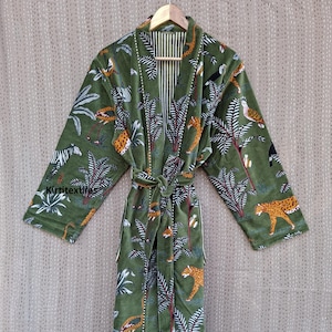 Puede incluir: Bata de terciopelo verde con un estampado de jungla con leopardos, cebras, pájaros y palmeras. La bata tiene un cinturón a juego y un forro interior a rayas. La bata está colgada de una percha de madera.