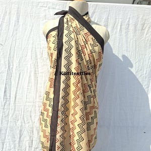 Könnte beinhalten: Ein beigefarbenes Sarong mit einem schwarzen und roten Zickzackmuster. Das Sarong ist auf einer Schaufensterpuppe drapiert, mit einem schwarzen Rand und dem Text "Kirititextiles". Der Stoff ist leicht und für warmes Wetter geeignet.