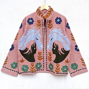 May include: A pink embroidered jacket with a geometric pattern. The jacket features blue horse heads, black silhouettes, and floral designs in blue, green, and brown. The jacket has a zipper closure and a high collar.