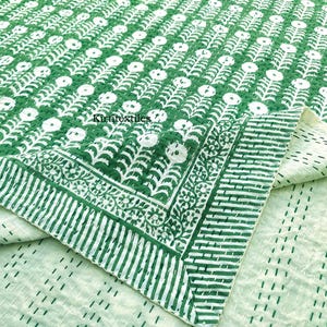 Puede incluir: Un edredón con estampado floral verde y blanco. El edredón presenta un diseño repetitivo de flores blancas y tallos verdes. El borde tiene un patrón de rayas. El edredón está doblado, revelando una parte inferior verde claro con líneas cosidas.