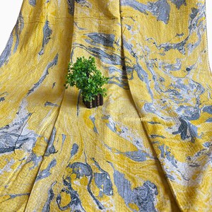 Op de afbeelding: Gele stof met een marmerpatroon in grijstinten en wit. De stof is gedrapeerd, waardoor zachte plooien en schaduwen ontstaan. Een kleine, kunstmatige groene plant ligt erop.