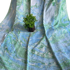 Könnte beinhalten: Ein blau, grün und weiß gemusterter Stoff mit marmorähnlichem Design. Der Stoff ist drapiert, und eine kleine Topfpflanze liegt obenauf. Der Stoff hat eine genähte Textur.
