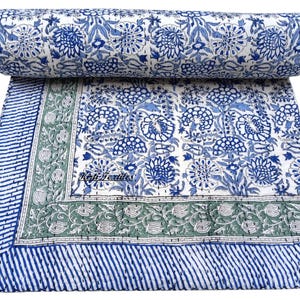 Könnte beinhalten: Eine weiße Baumwolldecke mit einem blauen und grünen Blumenmuster. Die Decke ist in der Hälfte gefaltet und hat einen Rand aus blauen und weißen Streifen.