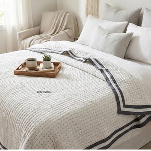 Op de afbeelding: Een wit gewatteerd sprei met een donkergrijze rand en bijpassende kussenset. Een houten dienblad bevat een koffiemok en een kleine potplant. Het bed is opgemaakt met witte lakens en kussens.