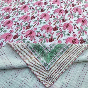 Op de afbeelding: Een witte quilt met een roze olifantenpatroon, palmbomen en aardbeien. De quilt heeft een groen-witte rand en is opgevouwen om een contrasterende witte onderkant met rode stiksels te laten zien. Het ontwerp combineert bloemen- en diermotieven.