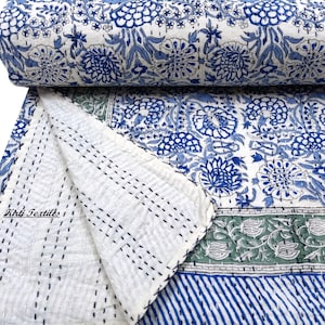Op de afbeelding: Een wit katoenen dekbed met een blauwe en groene bloemenprint. Het dekbed is met de hand gestikt en heeft een rand van blauwe strepen. Het dekbed is dubbelgevouwen en het label "Kirti Textiles" is zichtbaar.