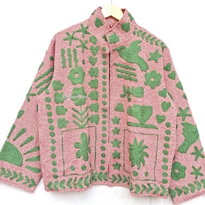 May include: A long-sleeved jacket in a dusty pink hue featuring a geometric diamond pattern throughout. The garment is adorned with raised green embroidery depicting various motifs including flowers, birds, hearts, stars, and a sun.