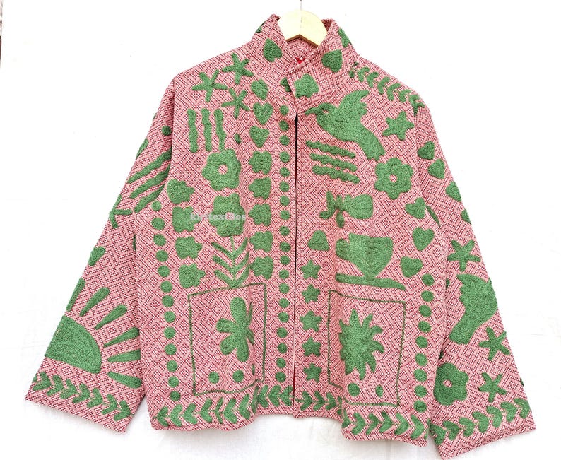 May include: A long-sleeved jacket in a dusty pink hue featuring a geometric diamond pattern throughout. The garment is adorned with raised green embroidery depicting various motifs including flowers, birds, hearts, stars, and a sun.