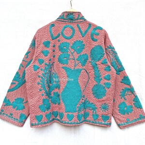 May include: A long-sleeved jacket in a dusty pink hue with a geometric diamond pattern, featuring vibrant teal embroidery. The back displays the word LOVE above a large pitcher filled with flowers, surrounded by heart and leaf motifs.