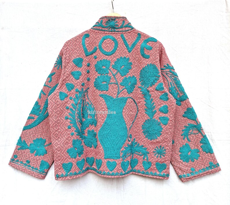 May include: A long-sleeved jacket in a dusty pink hue with a geometric diamond pattern, featuring vibrant teal embroidery. The back displays the word LOVE above a large pitcher filled with flowers, surrounded by heart and leaf motifs.
