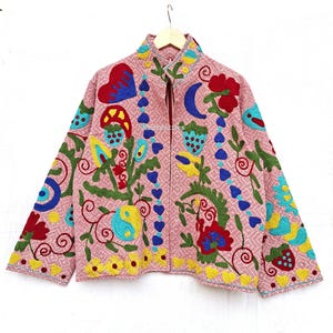 Könnte beinhalten: Eine rosa bestickte Jacke mit hohem Kragen und Reißverschluss. Die Jacke hat bunte Blumen- und abstrakte Designs in Rot, Blau, Gelb und Grün. Die Jacke hängt an einem Holzbügel.