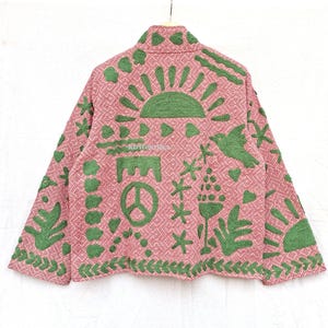 May include: A long-sleeved jacket in a dusty pink hue featuring a repeating geometric diamond pattern. The garment is adorned with raised green textured appliques depicting a sun, a peace sign, a bird, hearts, and various botanical motifs.