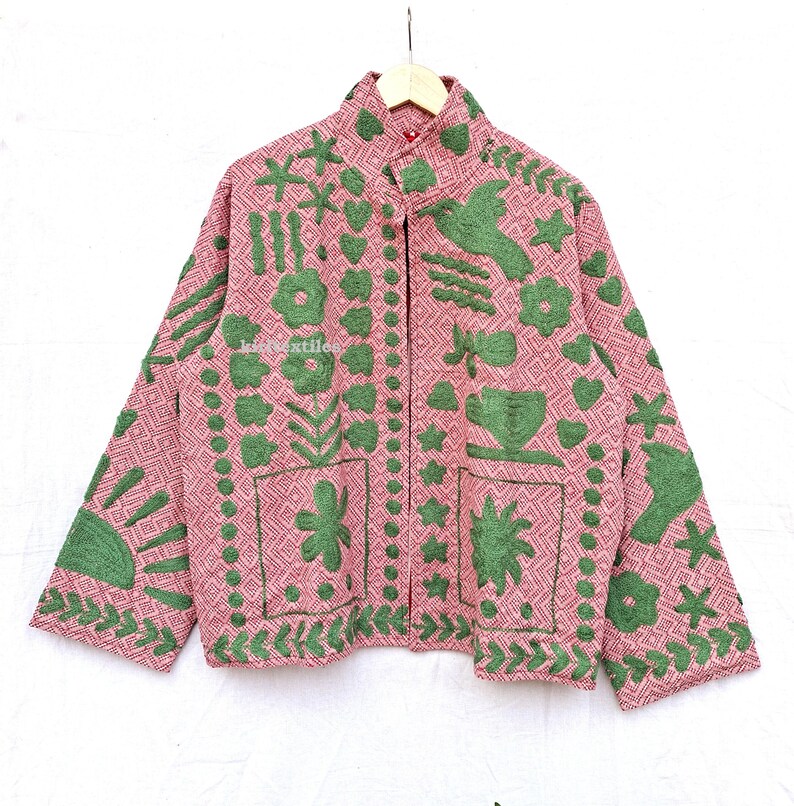 May include: A long-sleeved jacket in a dusty pink hue featuring a geometric diamond pattern and raised green embroidery. The design includes motifs of flowers, birds, stars, and a sun. The text "kidtextiles" is visible on the front panel.