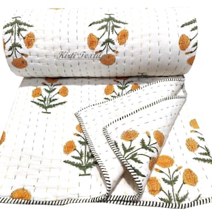 Könnte beinhalten: Weiße Baumwolldecke mit einem floralen Muster aus gelben Ringelblumen und grünen Blättern. Die Decke ist handgenäht und hat einen weißen Hintergrund mit einer subtilen Textur. Die Decke ist in der Hälfte gefaltet und hat einen schwarz-weißen Saum.