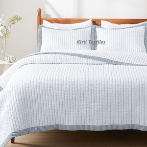 Könnte beinhalten: Ein weißer gesteppter Bettüberwurf mit einem blau-weiß gestreiften Rand. Der Bettüberwurf liegt auf einem Bett mit zwei weißen Kissen mit blau-weiß gestreiften Rändern. Der Text "Kirti Textiles" ist auf den Kissen sichtbar.