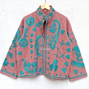 May include: A long-sleeved jacket featuring a pink and white geometric diamond pattern base with vibrant teal embroidered floral and heart motifs. The jacket has a high collar, a front zipper closure, and a structured fit with long sleeves.