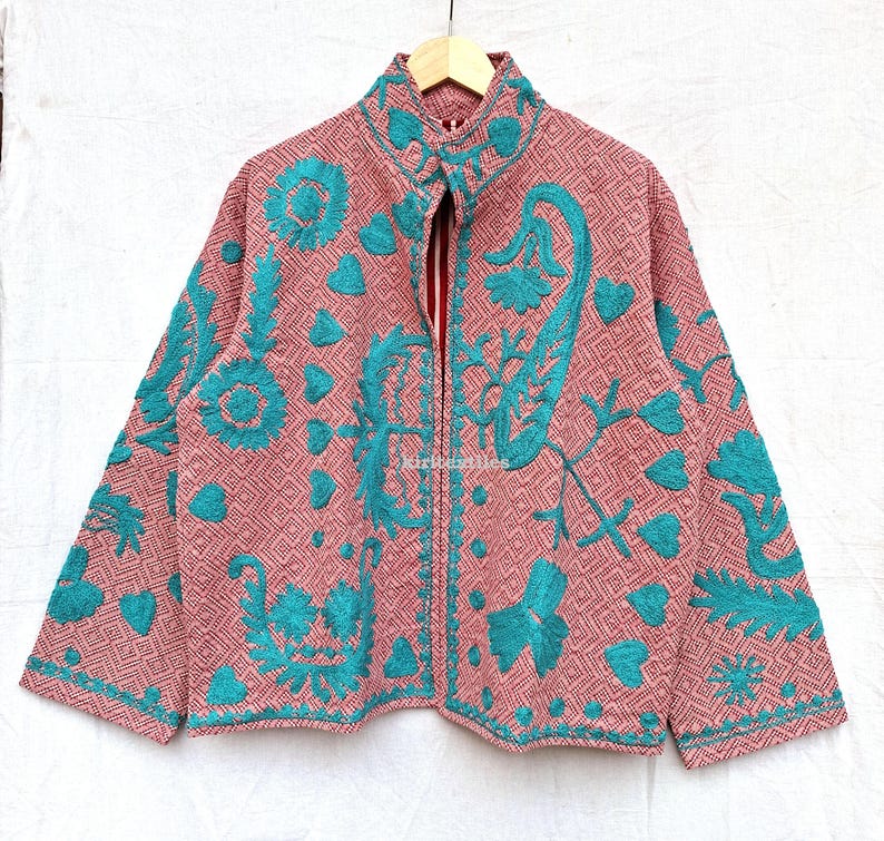 May include: A long-sleeved jacket featuring a pink and white geometric diamond pattern base with vibrant teal embroidered floral and heart motifs. The jacket has a high collar, a front zipper closure, and a structured fit with long sleeves.