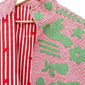 May include: A jacket featuring a red and white geometric pattern on the exterior with green textured appliqu&eacute;s of a bird, flower, and stars. The interior lining is red and white striped fabric with small red heart accents and the text kiritextiles.