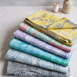 Könnte beinhalten: Ein Stapel aus fünf gefalteten Kantha-Steppdecken mit verschiedenen Marmormustern in Gelb, Rosa, Lavendel, Türkis und Grau. Jede Decke hat sichtbare weiße Steppstich-Stickereien und ein kleines weißes Stoffetikett mit der Aufschrift KIRTI TEXTILES.
